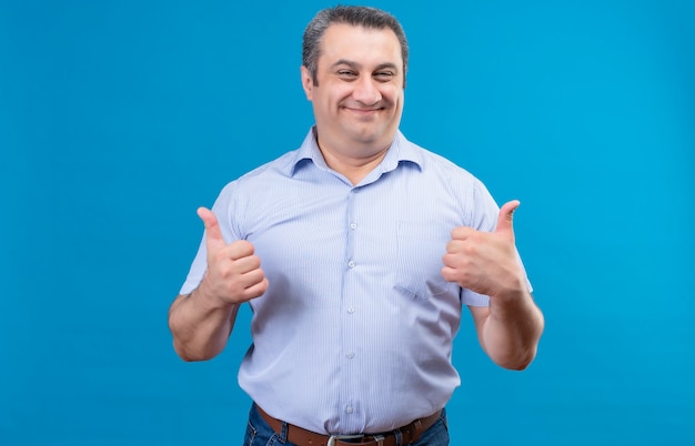 Homem de meia-idade feliz e animado com uma camisa azul, mostrando as duas mãos com o polegar para cima como um gesto em um espaço azul