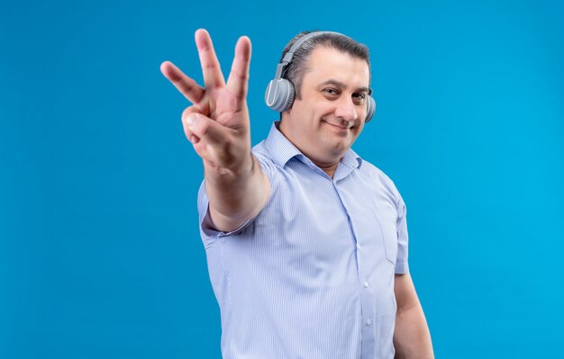 Homem de meia-idade feliz e positivo em uma camisa listrada azul em fones de ouvido, mostrando com os dedos número seis em um espaço azul