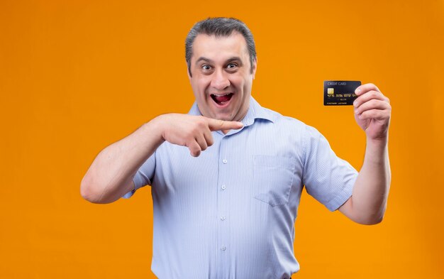 Homem de meia-idade feliz e sorridente, vestindo uma camisa azul listrada vertical apontando com o dedo indicador do cartão de crédito