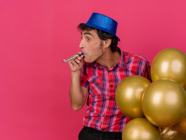 Homem de meia-idade impressionado com chapéu de festa e segurando balões, olhando para o lado soprando soprador de festa isolado na parede carmesim