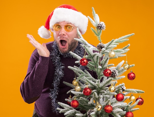 Homem de meia-idade impressionado com chapéu de papai noel e guirlanda de ouropel no pescoço, óculos em pé atrás de uma árvore de natal decorada, mostrando a mão vazia isolada na parede laranja