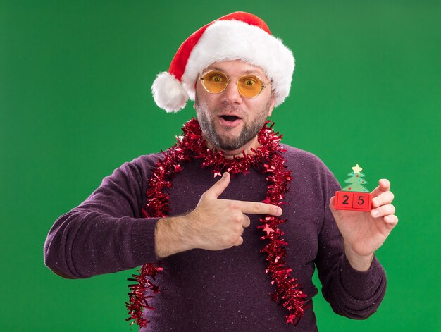 Homem de meia-idade impressionado com chapéu de Papai Noel e guirlanda de ouropel no pescoço, óculos segurando e apontando para a árvore de Natal, brinquedo com data, olhando para a câmera