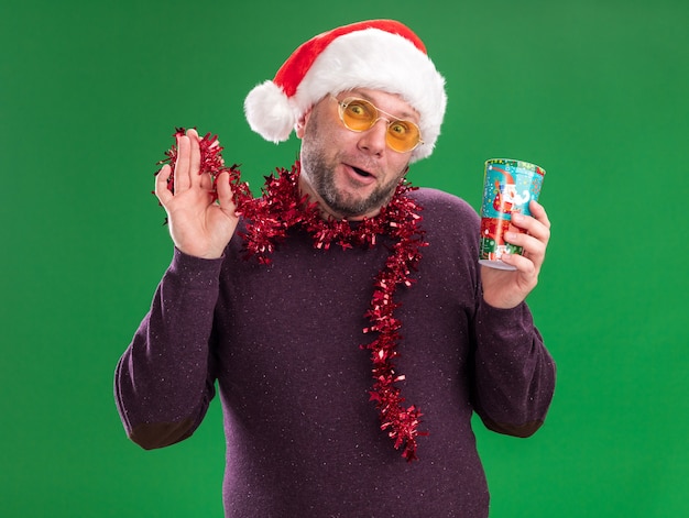 Foto grátis homem de meia-idade impressionado com chapéu de papai noel e guirlanda de ouropel no pescoço, óculos segurando um copo de natal de plástico segurando uma guirlanda de ouropel olhando para a câmera isolada sobre fundo verde