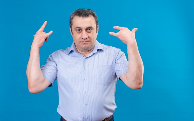 Homem de meia-idade problemático em uma camisa listrada azul de mãos dadas no símbolo da rocha sobre um fundo azul