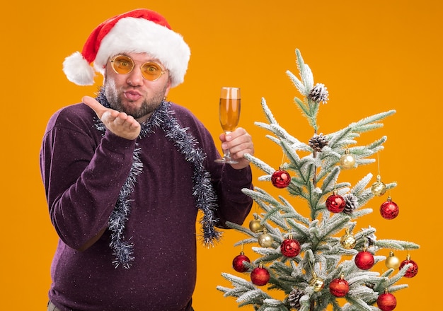 Homem de meia-idade satisfeito com chapéu de papai noel e guirlanda de ouropel no pescoço, óculos em pé perto da árvore de natal decorada segurando uma taça de champanhe