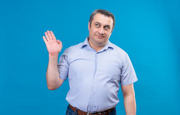 Homem de meia-idade satisfeito, vestindo uma camisa listrada azul, levantando a mão sobre um fundo azul