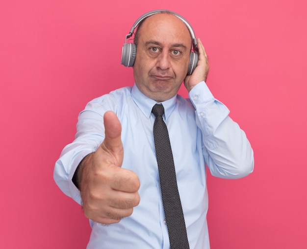Homem de meia-idade satisfeito, vestindo uma camiseta branca com gravata e fones de ouvido, mostrando o polegar isolado na parede rosa