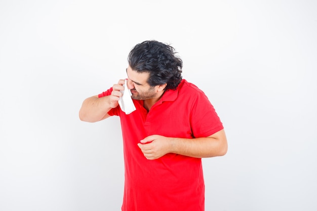 Homem de meia idade segurando o lenço assoando o nariz escorrendo em t-shirt vermelha e olhando insalubre, vista frontal.