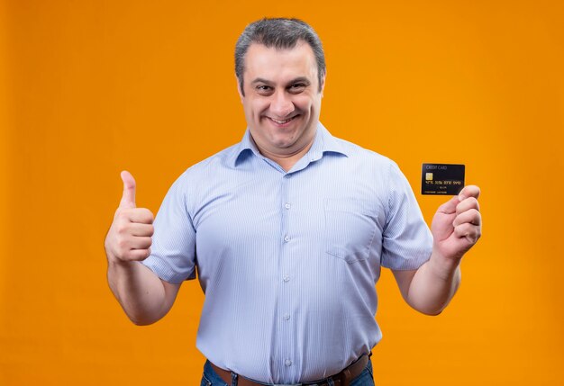 Homem de meia-idade sorridente e positivo com uma camisa listrada azul, segurando um cartão de crédito enquanto gesticula com o polegar para cima em um fundo laranja
