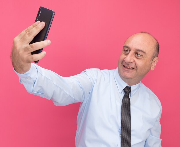 Homem de meia-idade sorridente usando camiseta branca com gravata tira uma selfie isolada na parede rosa