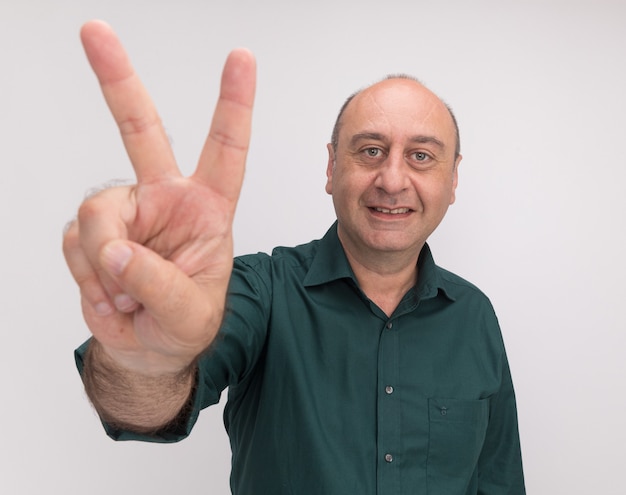 Foto grátis homem de meia-idade sorridente, vestindo uma camiseta verde, mostrando um gesto de paz isolado na parede branca