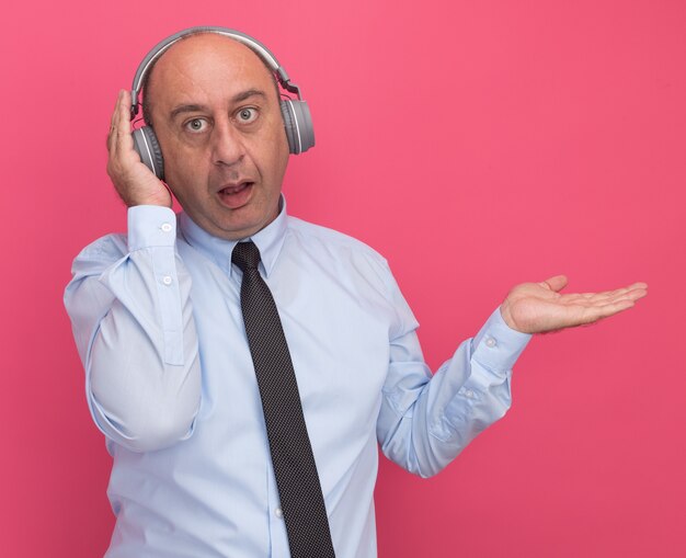 Homem de meia-idade surpreso vestindo camiseta branca com gravata e pontas de fones de ouvido com a mão ao lado isolada na parede rosa com espaço de cópia