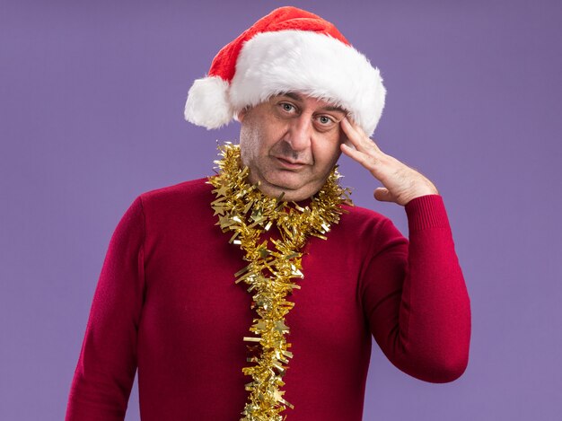 Homem de meia idade usando chapéu de Papai Noel de Natal com enfeites em volta do pescoço e olhando para a câmera confuso com uma mão na cabeça em pé sobre um fundo roxo