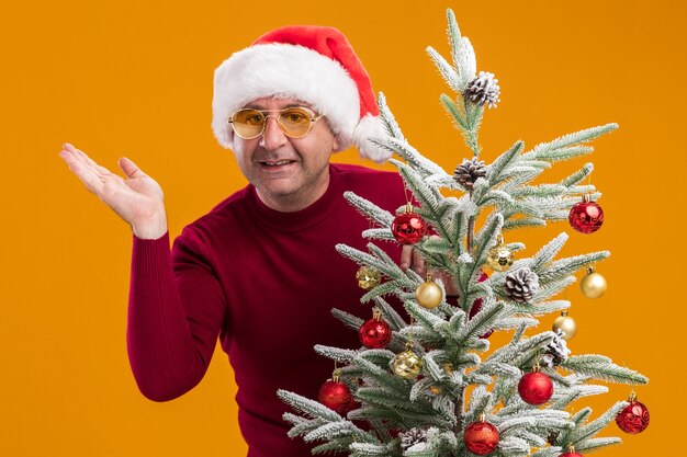 Homem de meia-idade usando chapéu de Papai Noel de Natal com gola alta e óculos amarelos, decorando a árvore de Natal, olhando para a câmera, sorrindo alegremente em pé sobre um fundo laranja