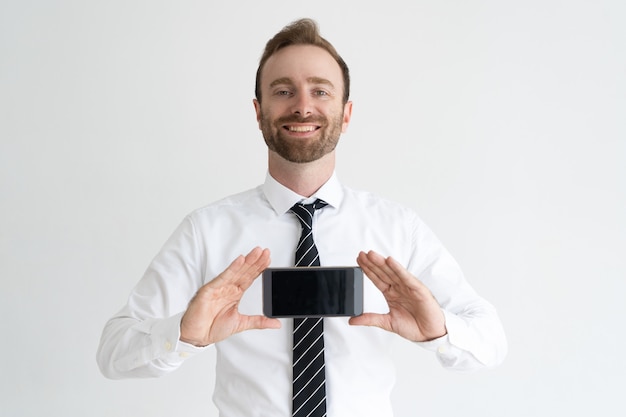 Homem de negócio de sorriso que guarda o smartphone, mostrando sua tela e olhando a câmera.