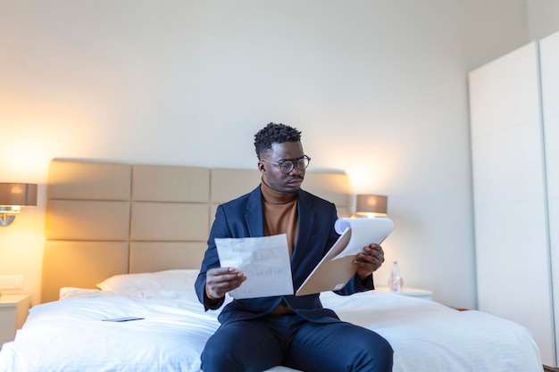 Homem de negócios aricano sério sentado na cama do hotel trabalhando analisa despesas em viagem de negócios no quarto de hotel