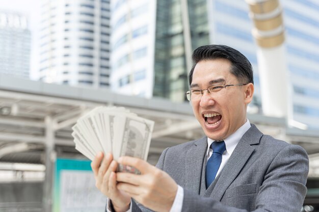 Homem de negócios asiático com cara feliz segurando notas de dólar americano no distrito comercial urbano