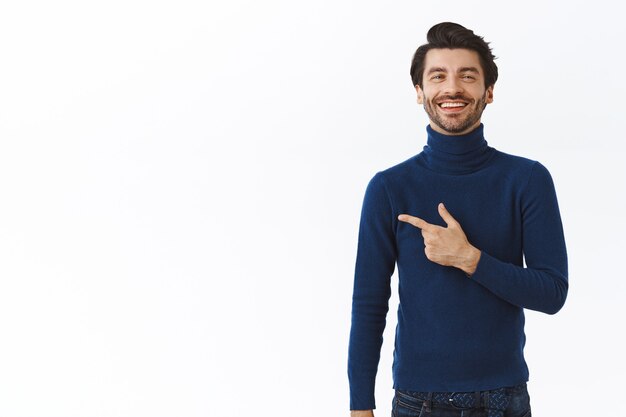 Homem de negócios barbudo, orgulhoso e bem-sucedido, em um suéter azul elegante de gola alta, apontando para a esquerda e sorrindo com expressão de satisfação, rindo enquanto se gabava de comprar um carro novo, parede branca