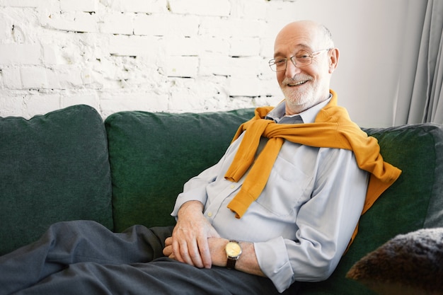 Homem de negócios bem-sucedido e atraente idoso usando óculos, camisa formal e moletom em volta do pescoço, sentado no sofá de seu escritório, com um sorriso radiante, feliz depois de fazer um bom negócio