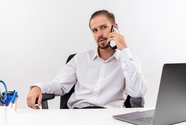 Homem de negócios bonito com camisa branca e fones de ouvido com microfone olhando para o lado com cara séria falando no celular, sentado à mesa em escritório sobre fundo branco