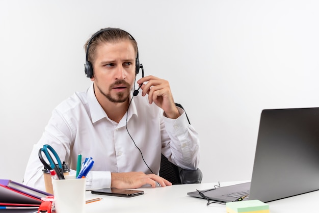 Homem de negócios bonito com camisa branca e fones de ouvido com microfone ouvindo um cliente com cara séria sentado à mesa em escritório sobre fundo branco