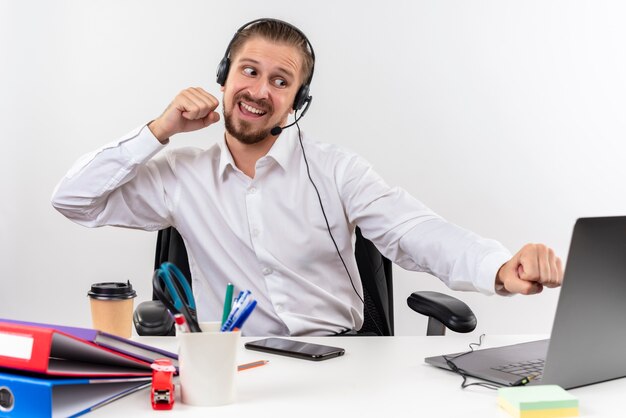 Homem de negócios bonito com camisa branca e fones de ouvido com um microfone de punho cerrado muito zangado sentado à mesa em escritório sobre fundo branco