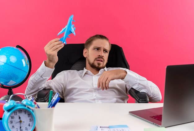 Homem de negócios bonito com camisa branca segurando um avião de brinquedo olhando de lado perplexo, sentado à mesa em escritório sobre fundo rosa