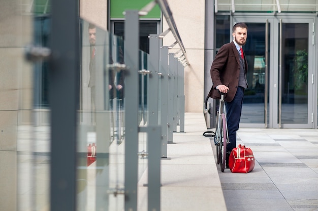 Homem de negócios bonito com uma jaqueta e gravata vermelha e sua bicicleta nas ruas da cidade. a bolsa vermelha está a seguir. O conceito de estilo de vida moderno dos jovens