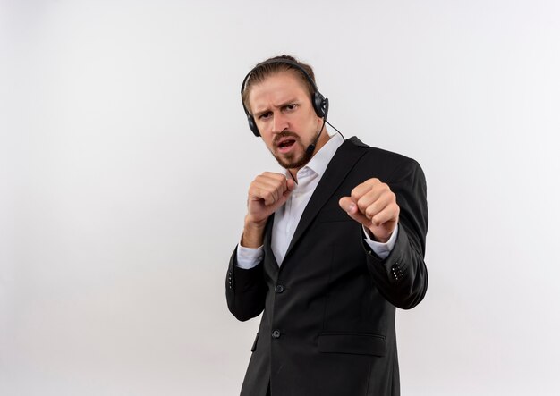 Homem de negócios bonito de terno e fones de ouvido com microfone, olhando para a câmera com cara séria, posando como um boxeador em pé sobre um fundo branco