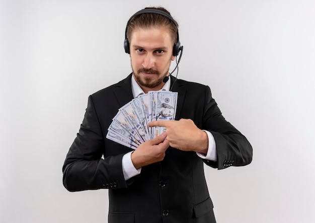 Homem de negócios bonito de terno e fones de ouvido com um microfone mostrando dinheiro apontando com o dedo para ele, olhando para a câmera com um sorriso no rosto em pé sobre um fundo branco