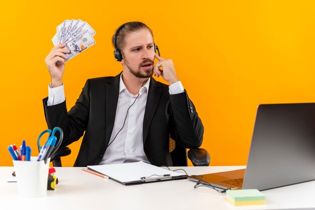 Homem de negócios bonito de terno e fones de ouvido com um microfone mostrando dinheiro olhando de lado pensando sentado à mesa em escritório sobre fundo laranja