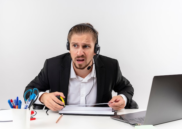 Homem de negócios bonito de terno e fones de ouvido com um microfone, olhando de lado e confuso, sentado à mesa em escritório sobre fundo branco