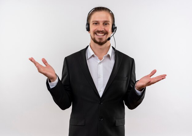 Homem de negócios bonito de terno e fones de ouvido com um microfone olhando para a câmera feliz e positivo sorrindo alegremente em pé sobre um fundo branco