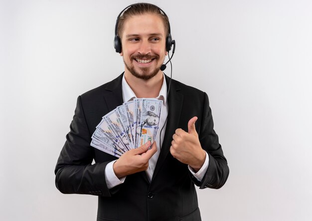 Homem de negócios bonito de terno e fones de ouvido com um microfone, olhando para a câmera, mostrando o dinheiro sorrindo alegremente mostrando os polegares em pé sobre um fundo branco