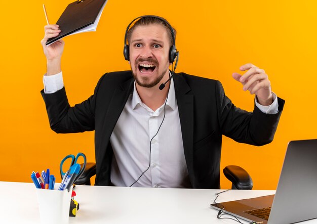 Homem de negócios bonito de terno e fones de ouvido com um microfone segurando a prancheta, olhando para o lado, gritando com uma expressão agressiva, sentado à mesa em escritório sobre fundo laranja