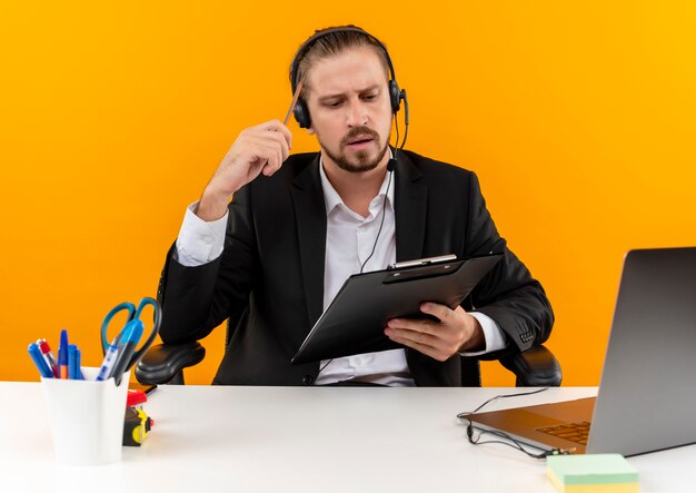 Homem de negócios bonito de terno e fones de ouvido com um microfone segurando uma prancheta, olhando para ela com uma cara séria, sentado à mesa em escritório sobre fundo laranja