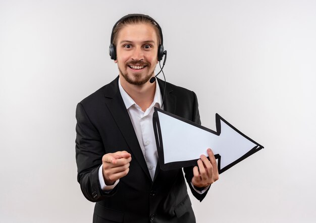 Homem de negócios bonito de terno e fones de ouvido com um microfone segurando uma seta branca apontando com o dedo para a câmera, sorrindo em pé sobre um fundo branco