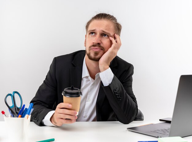 Homem de negócios bonito de terno segurando a xícara de café, trabalhando no laptop, olhando de lado cansado e entediado sentado à mesa em escritório sobre fundo branco