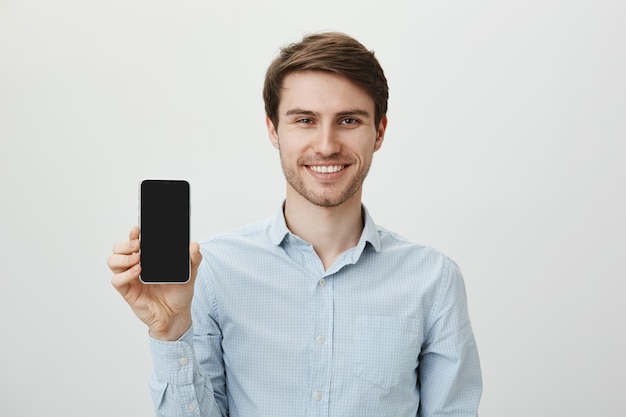 Foto grátis homem de negócios bonito e sorridente promove aplicativo móvel, exibindo tela de smartphone