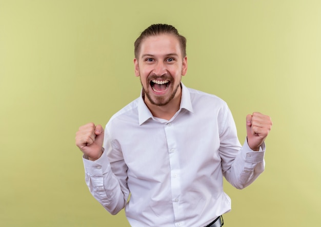 Homem de negócios bonito em camisa branca cerrando os punhos feliz e animado regozijando-se com seu sucesso em pé sobre fundo verde-oliva