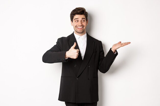 Homem de negócios bonito em terno preto, mostrando o polegar para cima e segurando seu produto na mão sobre o espaço branco da cópia, de pé contra um fundo branco.