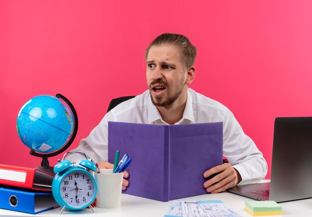 Homem de negócios bonito em uma camisa branca com um globo segurando um caderno, olhando de lado e confuso, sentado à mesa em escritório sobre fundo rosa