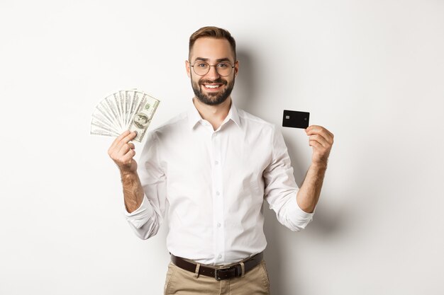 Homem de negócios bonito mostrando cartão de crédito e dólares, sorrindo satisfeito, em pé