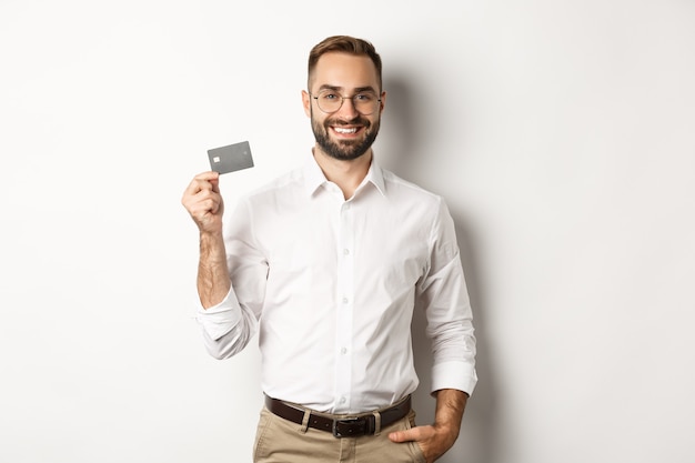 Homem de negócios bonito mostrando seu cartão de crédito, parecendo satisfeito, de pé