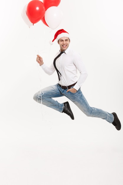 Homem de negócios bonito pulando para comemorar o feliz Natal, usando chapéu de Papai Noel com balão vermelho.