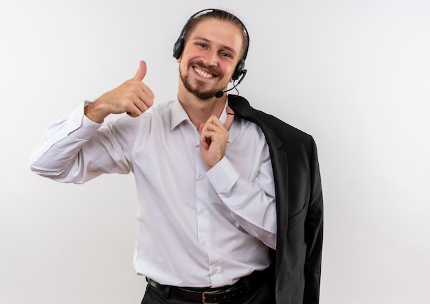 Homem de negócios bonito segurando uma jaqueta por cima do ombro com fones de ouvido e um microfone olhando para a câmera com um sorriso no rosto mostrando os polegares em pé sobre um fundo branco