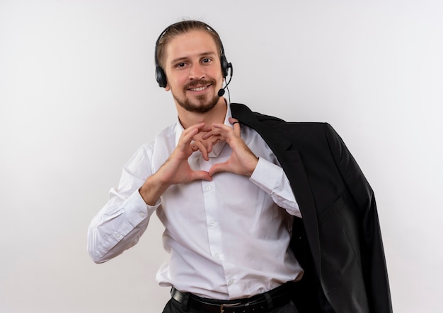 Foto grátis homem de negócios bonito segurando uma jaqueta sobre o ombro com fones de ouvido e um microfone fazendo um gesto de coração com os dedos sorrindo em pé sobre um fundo branco