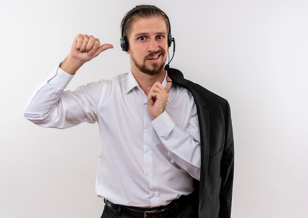 Homem de negócios bonito segurando uma jaqueta sobre o ombro com fones de ouvido e um microfone olhando para a câmera confuso em pé sobre um fundo branco