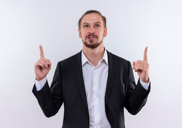 Homem de negócios bonito usando terno apontando para cima com os dedos indicadores, feliz e positivo, sorrindo em pé sobre um fundo branco