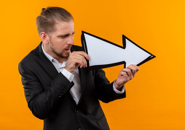 Homem de negócios bonito usando terno segurando uma seta branca apontando para o lado, parecendo confuso em pé sobre um fundo laranja